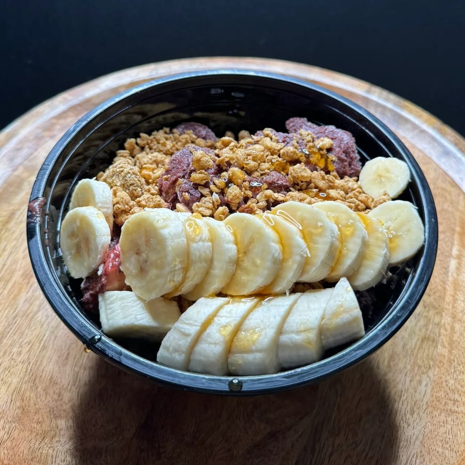 A Da Kane's Kitchen acai bowl with granola, banana slices, and honey drizzle in a black bowl.
